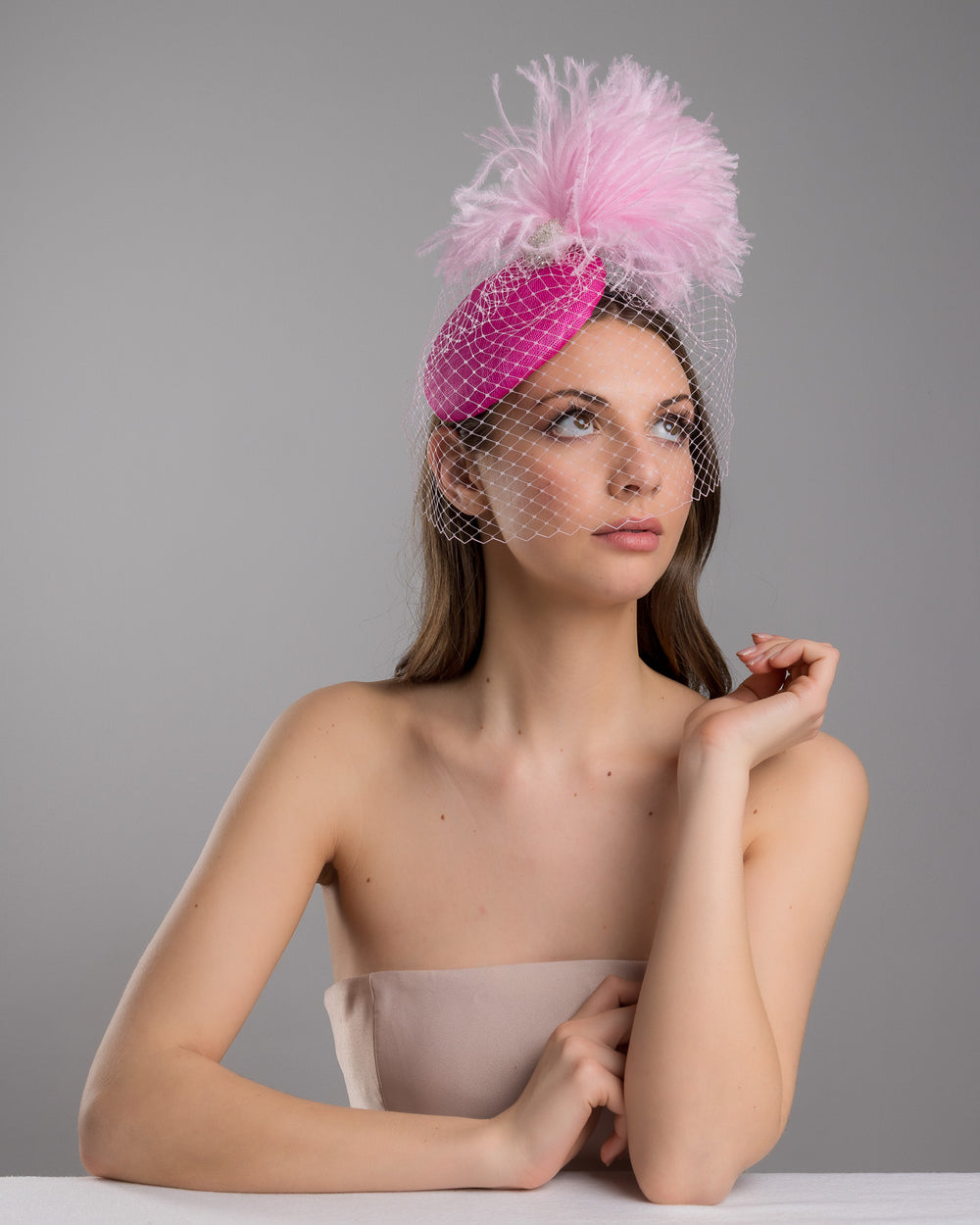 The female model is posing, wearing
Ever, the fuchsia velvet padded headband with a pink feather pom-pom detail, finished with a soft birdcage veil overlay and mounted on a slim black headband. Designed and made by Merve Bayindir Millinery.
