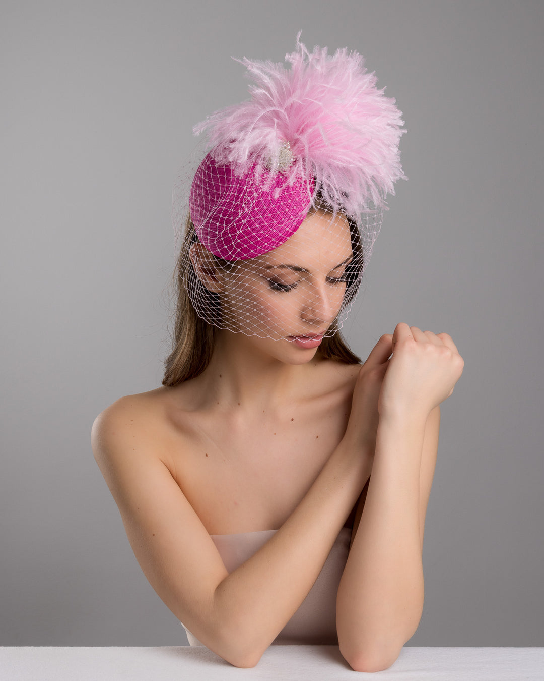 The female model is posing, wearing
Ever, the fuchsia velvet padded headband with a pink feather pom-pom detail, finished with a soft birdcage veil overlay and mounted on a slim black headband. Designed and made by Merve Bayindir Millinery.
