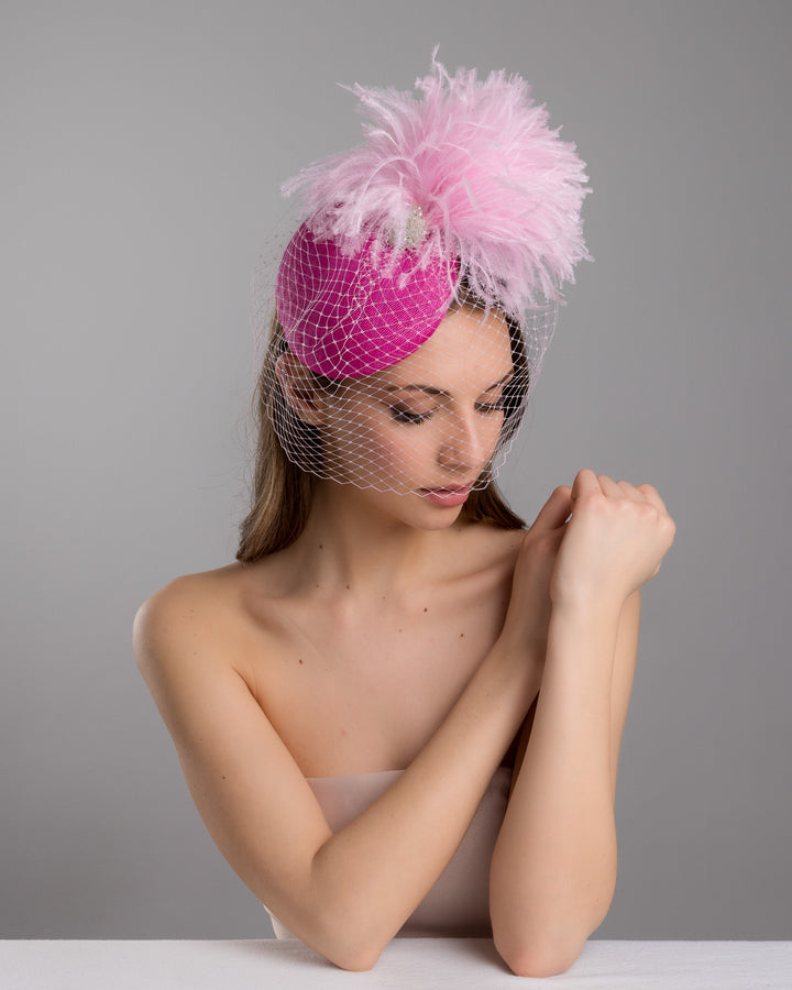The female model is posing, wearing
Ever, the fuchsia velvet padded headband with a pink feather pom-pom detail, finished with a soft birdcage veil overlay and mounted on a slim black headband. Designed and made by Merve Bayindir Millinery.
