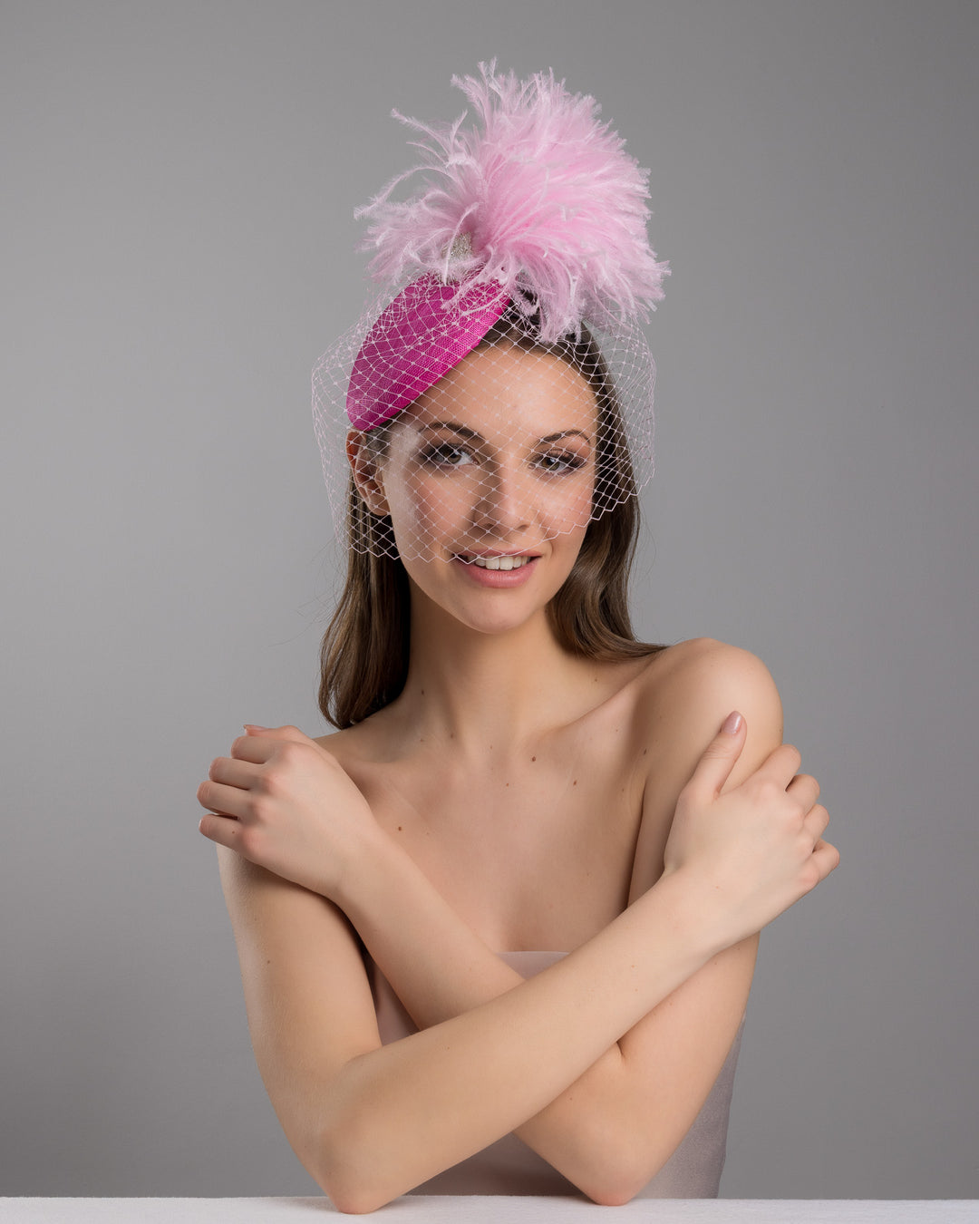 The female model is posing, wearing
Ever, the fuchsia velvet padded headband with a pink feather pom-pom detail, finished with a soft birdcage veil overlay and mounted on a slim black headband. Designed and made by Merve Bayindir Millinery.
