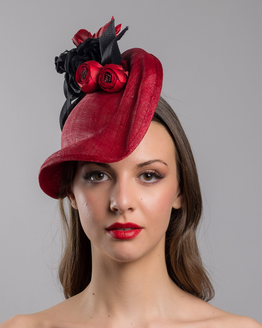 Model is showcasing the front side close up of asymmetric, curved red hat worn to the right side of the head. This headpiece is decorated with a bouquet of black and red crepe roses, black shaded red peonies and black buntal swirls that create a strong contrast and bond between each element. Designed and made by Merve Bayindir Millinery.