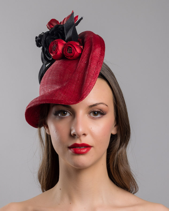 Model is showcasing the front side close up of asymmetric, curved red hat worn to the right side of the head. This headpiece is decorated with a bouquet of black and red crepe roses, black shaded red peonies and black buntal swirls that create a strong contrast and bond between each element. Designed and made by Merve Bayindir Millinery.