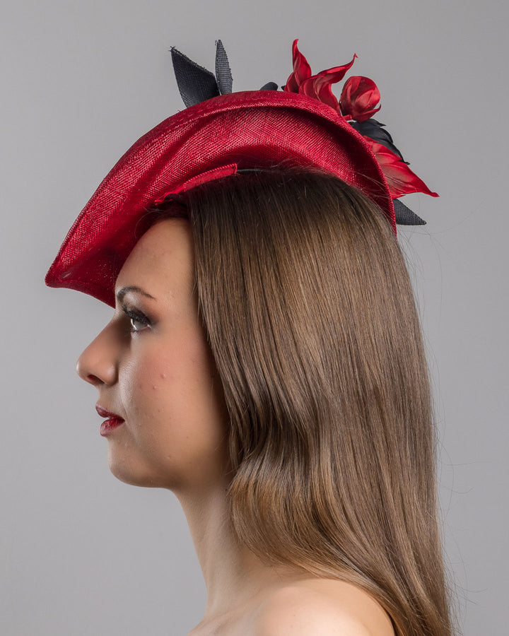 Model is showcasing the left side close up of asymmetric, curved red hat worn to the right side of the head. This headpiece is decorated with a bouquet of black and red crepe roses, black shaded red peonies and black buntal swirls that create a strong contrast and bond between each element. Designed and made by Merve Bayindir Millinery.