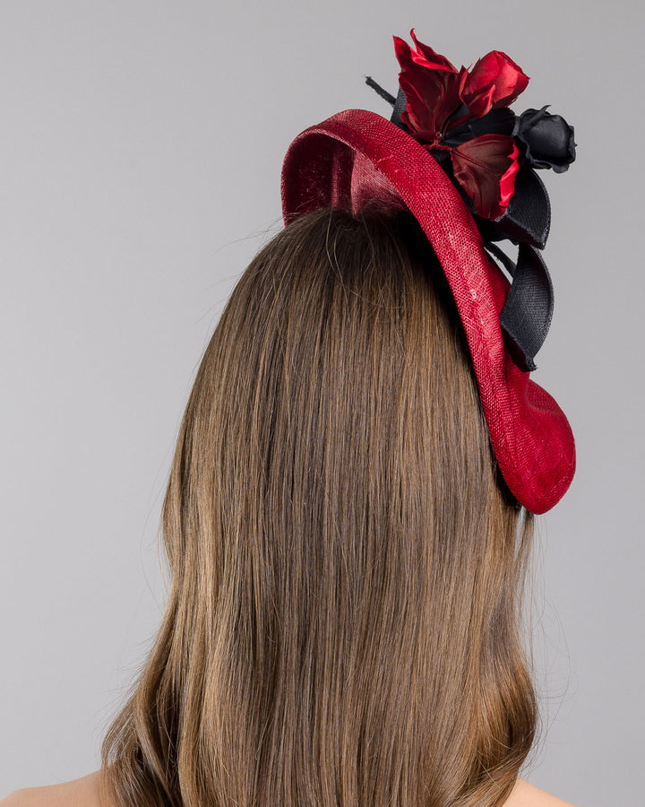 Model is showcasing the back side close up of asymmetric, curved red hat worn to the right side of the head. This headpiece is decorated with a bouquet of black and red crepe roses, black shaded red peonies and black buntal swirls that create a strong contrast and bond between each element. Designed and made by Merve Bayindir Millinery.