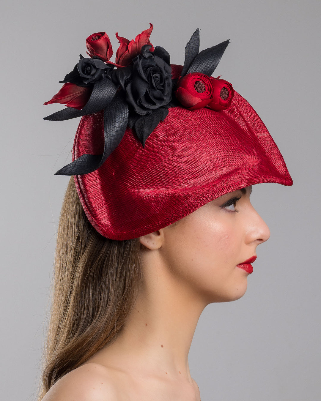 Model is showcasing the right side close up of asymmetric, curved red hat worn to the right side of the head. This headpiece is decorated with a bouquet of black and red crepe roses, black shaded red peonies and black buntal swirls that create a strong contrast and bond between each element. Designed and made by Merve Bayindir Millinery.
