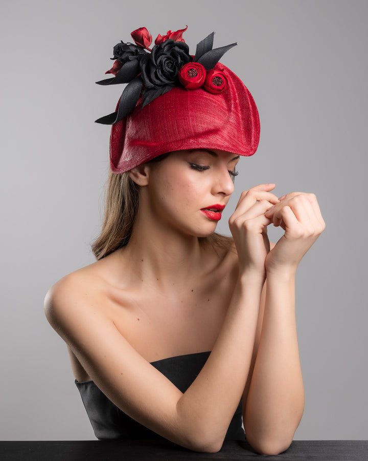 Model is posing wearing Echo the asymmetric, curved red hat worn to the right side of the head. This headpiece is decorated with a bouquet of black and red crepe roses, black shaded red peonies and black buntal swirls that create a strong contrast and bond between each element. Designed and made by Merve Bayindir Millinery.