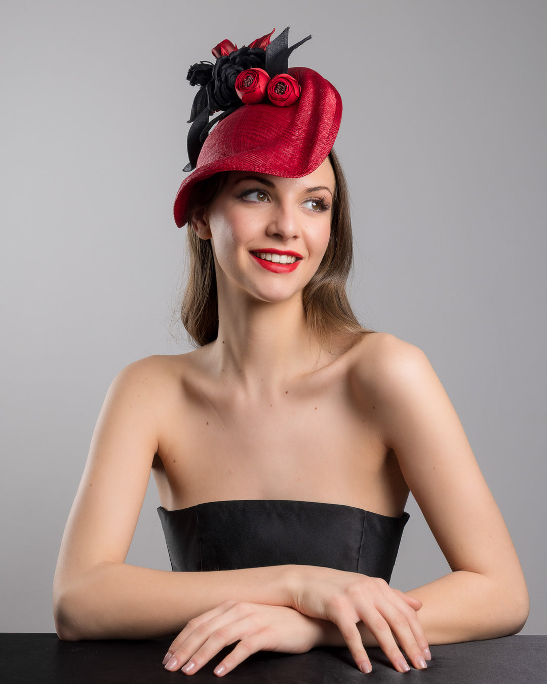Model is posing wearing Echo the asymmetric, curved red hat worn to the right side of the head. This headpiece is decorated with a bouquet of black and red crepe roses, black shaded red peonies and black buntal swirls that create a strong contrast and bond between each element. Designed and made by Merve Bayindir Millinery.