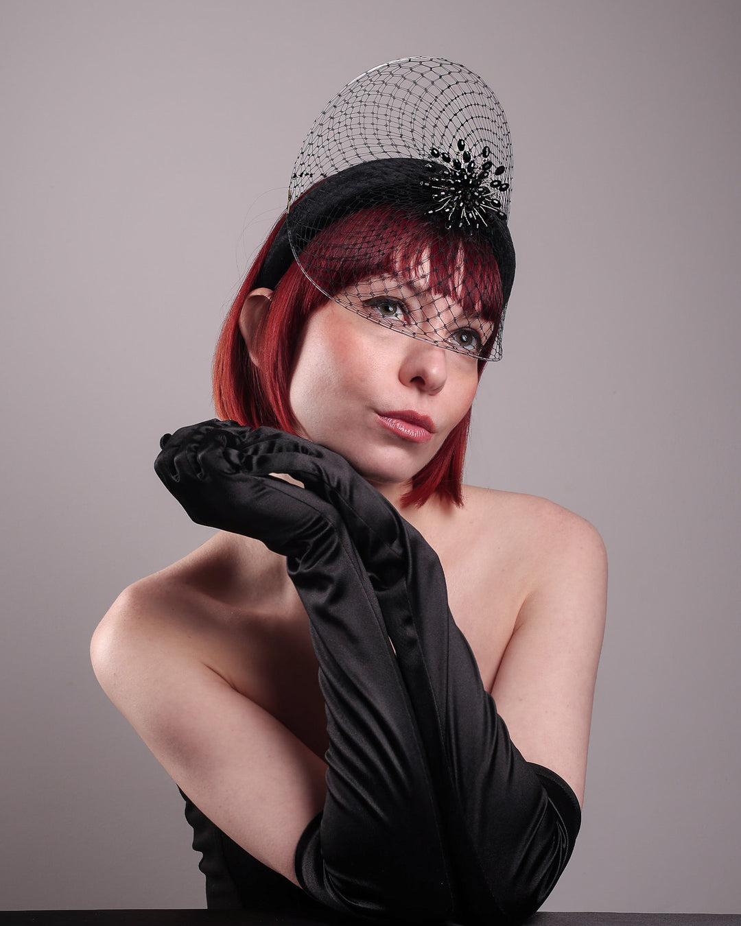The model showcases the low angle right front side of the Eclat headband veil mask, a tall sculptural headband form in black velvet is crowned by a jet-black beaded starburst and wrapped in delicate oval veiling that gracefully shields the eyes. Designed by Merve Bayindir.