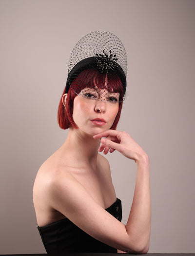 The model showcases the front of the Eclat headband veil mask, a tall sculptural headband form in black velvet is crowned by a jet-black beaded starburst and wrapped in delicate oval veiling that gracefully shields the eyes. Designed by Merve Bayindir.