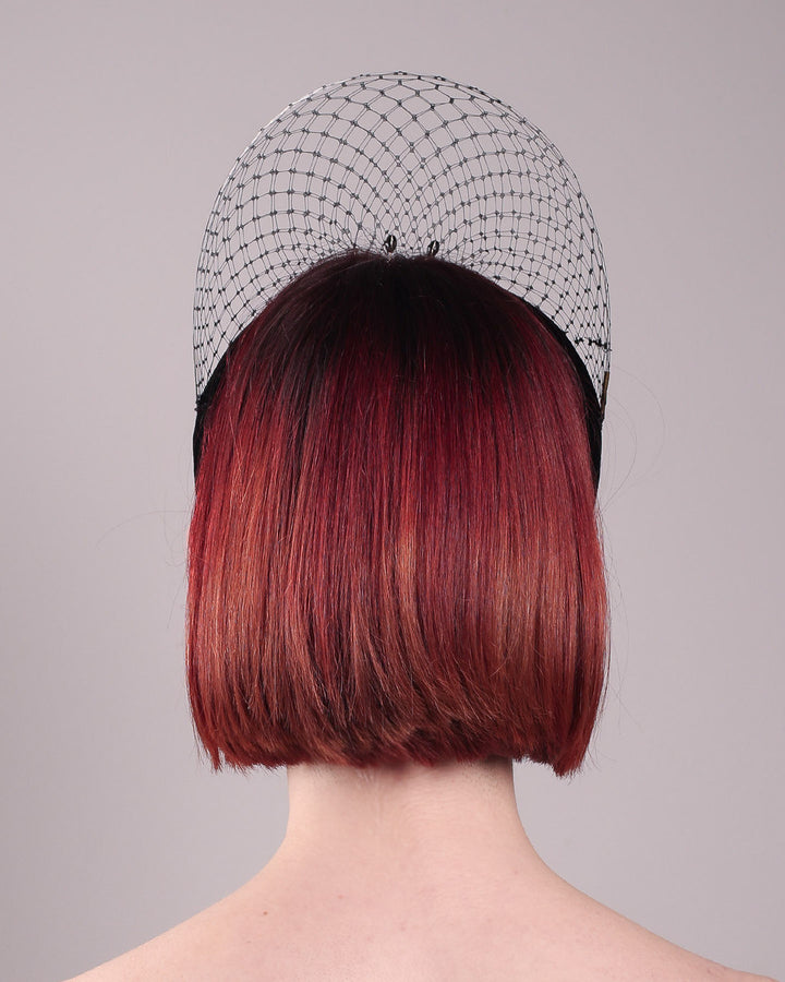 The model showcases the back of the Eclat headband veil mask, a tall sculptural headband form in black velvet is crowned by a jet-black beaded starburst and wrapped in delicate oval veiling that gracefully shields the eyes. Designed by Merve Bayindir.