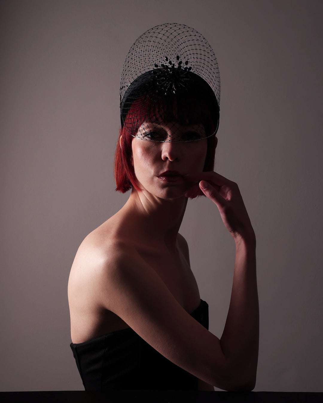 The model showcases un an edittorial lightining the front of the Eclat headband veil mask, a tall sculptural headband form in black velvet is crowned by a jet-black beaded starburst and wrapped in delicate oval veiling that gracefully shields the eyes. Designed by Merve Bayindir.