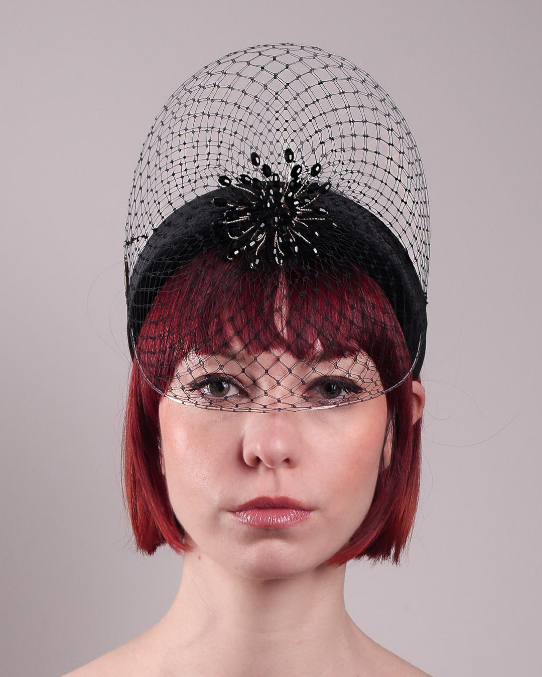 The model showcases the front close up of the Eclat headband veil mask, a tall sculptural headband form in black velvet is crowned by a jet-black beaded starburst and wrapped in delicate oval veiling that gracefully shields the eyes. Designed by Merve Bayindir.