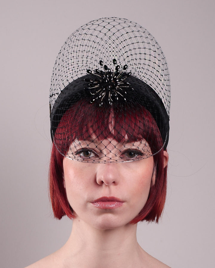 The model showcases the front close up of the Eclat headband veil mask, a tall sculptural headband form in black velvet is crowned by a jet-black beaded starburst and wrapped in delicate oval veiling that gracefully shields the eyes. Designed by Merve Bayindir.