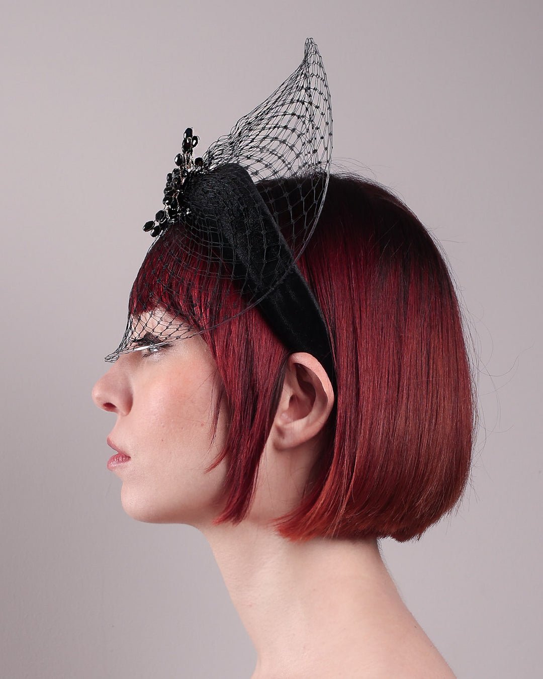 The model showcases the left close up of the Eclat headband veil mask, a tall sculptural headband form in black velvet is crowned by a jet-black beaded starburst and wrapped in delicate oval veiling that gracefully shields the eyes. Designed by Merve Bayindir.