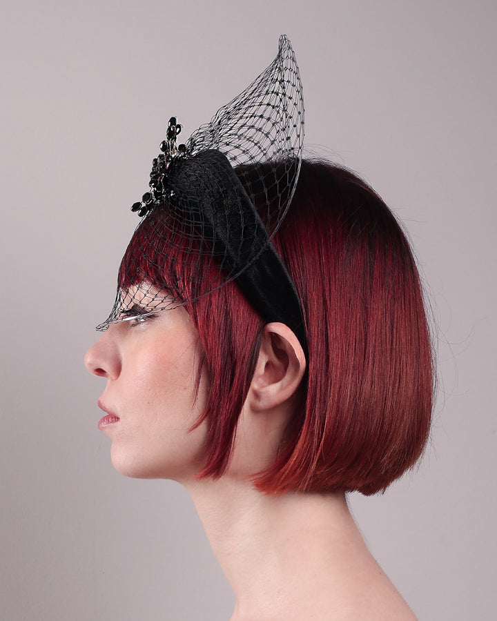The model showcases the left close up of the Eclat headband veil mask, a tall sculptural headband form in black velvet is crowned by a jet-black beaded starburst and wrapped in delicate oval veiling that gracefully shields the eyes. Designed by Merve Bayindir.