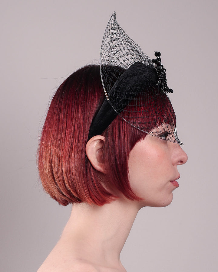 The model showcases the right side close up of the Eclat headband veil mask, a tall sculptural headband form in black velvet is crowned by a jet-black beaded starburst and wrapped in delicate oval veiling that gracefully shields the eyes. Designed by Merve Bayindir.