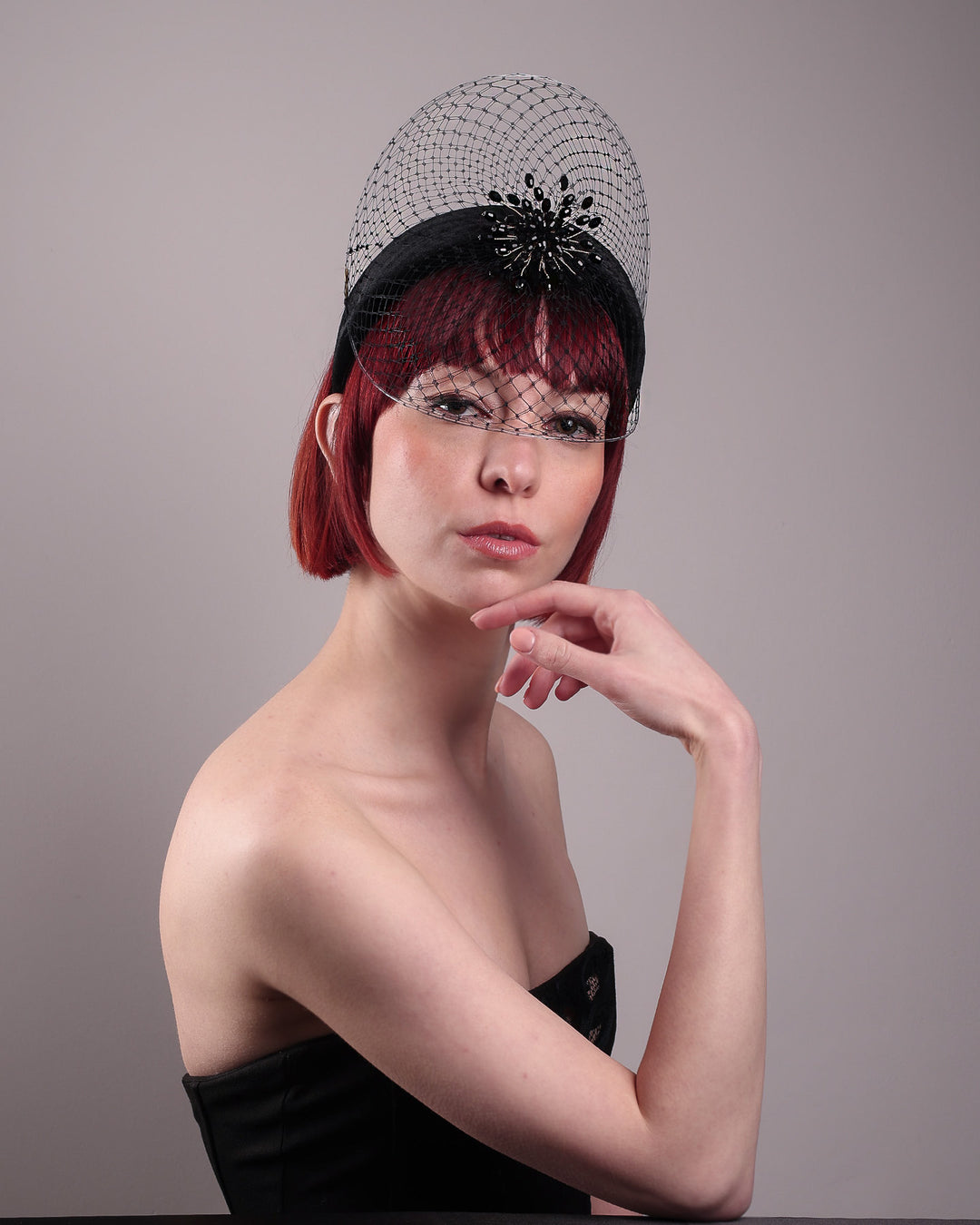 The model showcases the front of the Eclat headband veil mask, a tall sculptural headband form in black velvet is crowned by a jet-black beaded starburst and wrapped in delicate oval veiling that gracefully shields the eyes. Designed by Merve Bayindir.