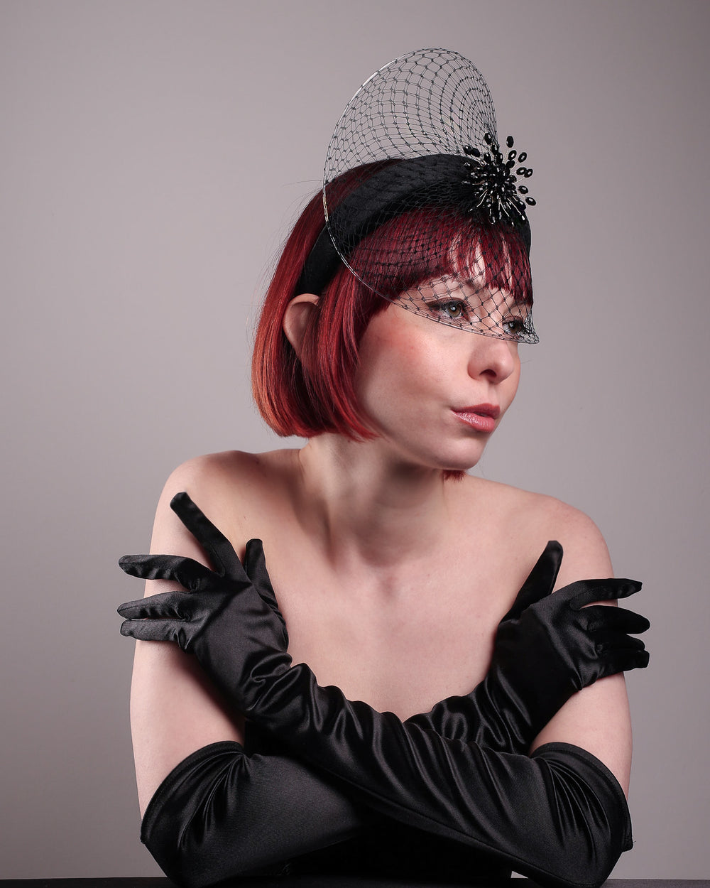 The model showcases the right front of the Eclat headband veil mask, a tall sculptural headband form in black velvet is crowned by a jet-black beaded starburst and wrapped in delicate oval veiling that gracefully shields the eyes. Designed by Merve Bayindir.