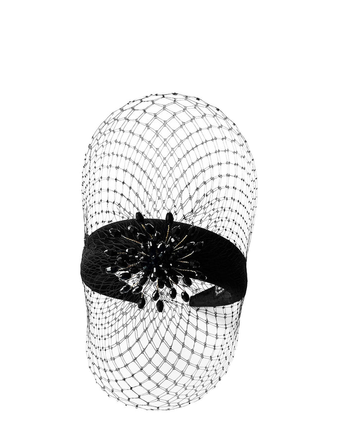 Still life picture of the Eclat headband veil mask, a tall sculptural headband form in black velvet is crowned by a jet-black beaded starburst and wrapped in delicate oval veiling that gracefully shields the eyes. Designed by Merve Bayindir.
