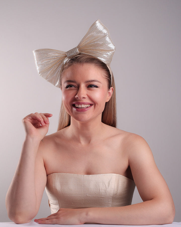 Model is showcasing the left front of a light beige/ivory alice style silk headband is decorated with a large bow with shimmering metallic bow placed on the right centre of the headband.
