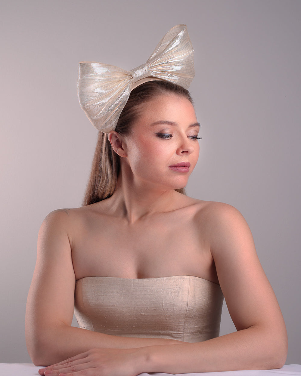Model is showcasing the right front angle of a light beige/ivory alice style silk headband is decorated with a large bow with shimmering metallic bow placed on the right centre of the headband.