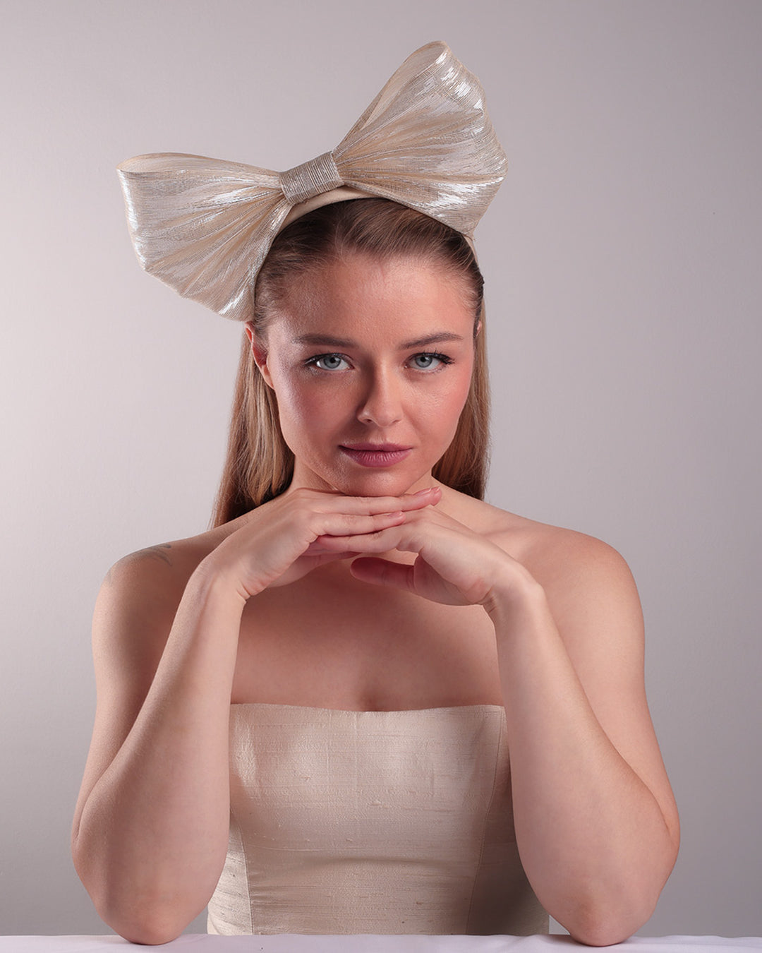 Model is showcasing the front of a light beige/ivory alice style silk headband is decorated with a large bow with shimmering metallic bow placed on the right centre of the headband.