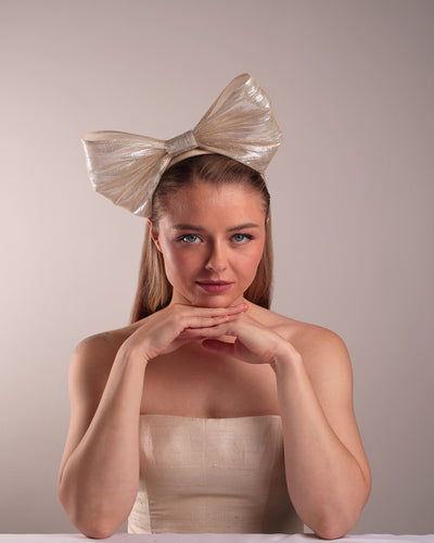 Model is showcasing the front of a light beige/ivory alice style silk headband is decorated with a large bow with shimmering metallic bow placed on the right centre of the headband.