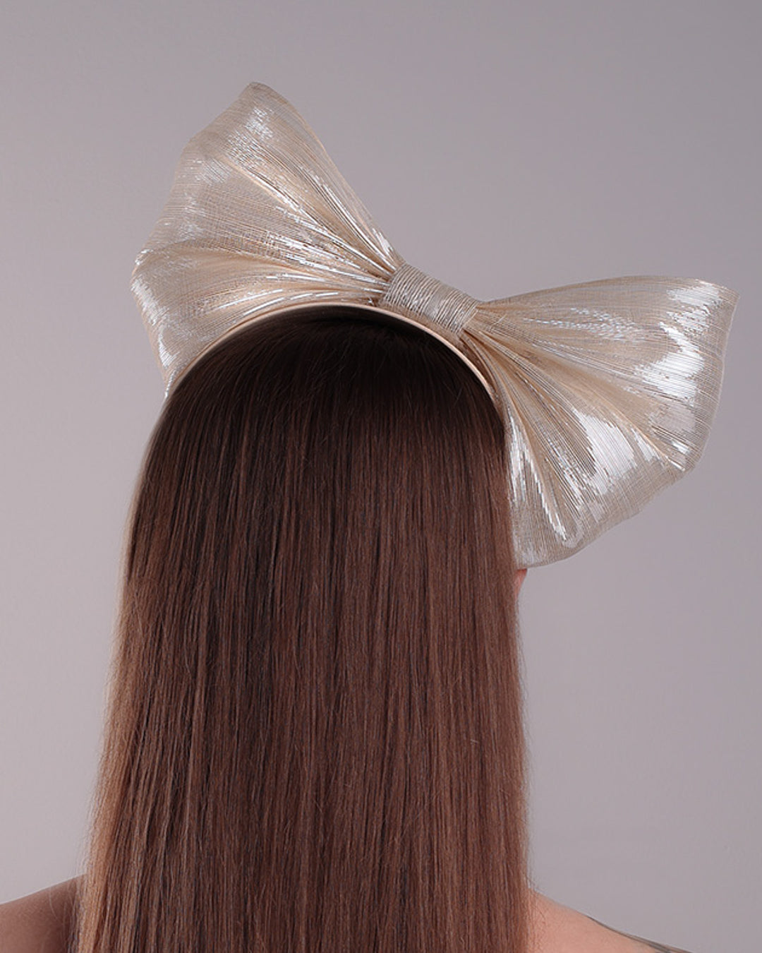 Model is showcasing the back of a light beige/ivory alice style silk headband is decorated with a large bow with shimmering metallic bow placed on the right centre of the headband.