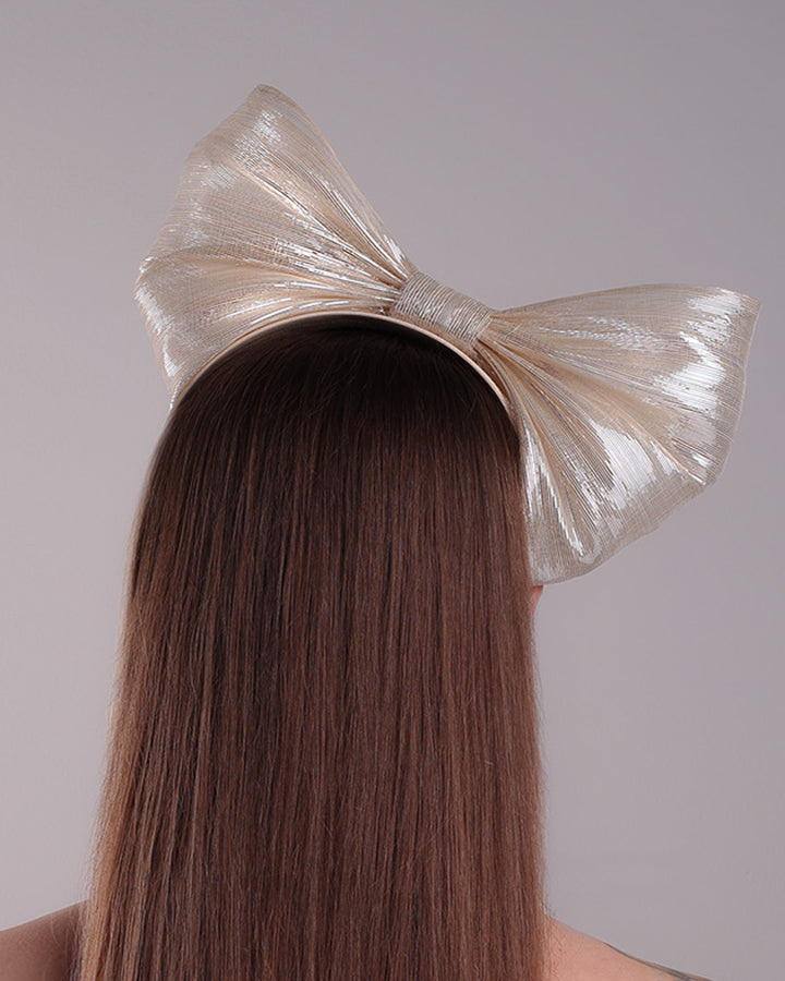 Model is showcasing the back of a light beige/ivory alice style silk headband is decorated with a large bow with shimmering metallic bow placed on the right centre of the headband.