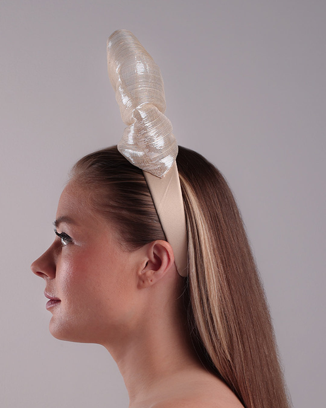 Model is showcasing the left side of a light beige/ivory alice style silk headband is decorated with a large bow with shimmering metallic bow placed on the right centre of the headband.