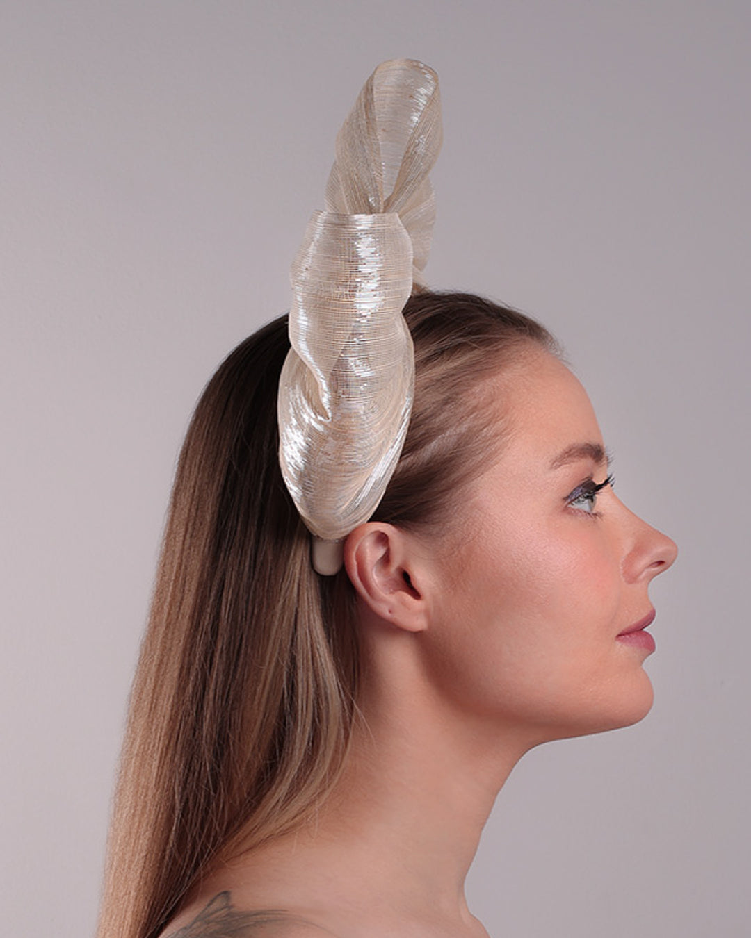 Model is showcasing the right side of a light beige/ivory alice style silk headband is decorated with a large bow with shimmering metallic bow placed on the right centre of the headband.