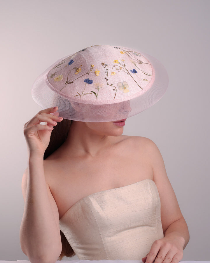 The model is showcasing the front right top side of the circle shaped pink sinamay saucer hat, worn tilted to the right side. The edges of the circle is embellised with natural pressed dried flowers. The entire cirlce hat extends with pink crinoline at the edge. The saucer hat is fitted with a wire headband and supported with elastic band.