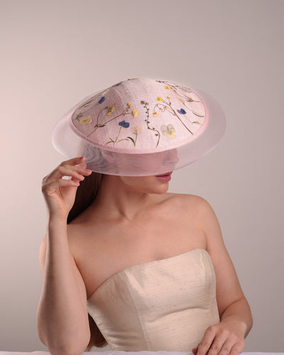 The model is showcasing the front right top side of the circle shaped pink sinamay saucer hat, worn tilted to the right side. The edges of the circle is embellised with natural pressed dried flowers. The entire cirlce hat extends with pink crinoline at the edge. The saucer hat is fitted with a wire headband and supported with elastic band.