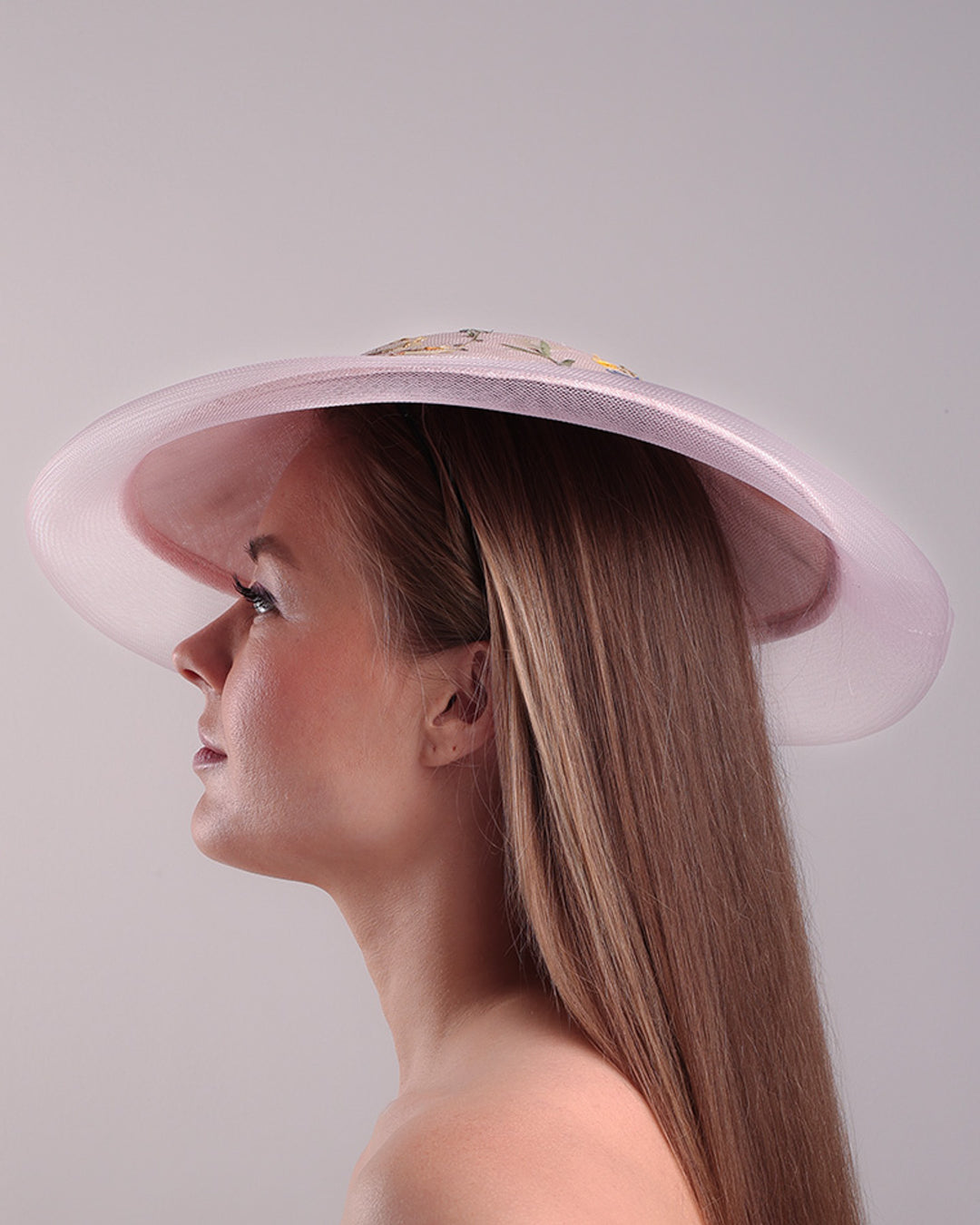 The model is showcasing the left side of the circle shaped pink sinamay saucer hat, worn tilted to the right side. The edges of the circle is embellised with natural pressed dried flowers. The entire cirlce hat extends with pink crinoline at the edge. The saucer hat is fitted with a wire headband and supported with elastic band.