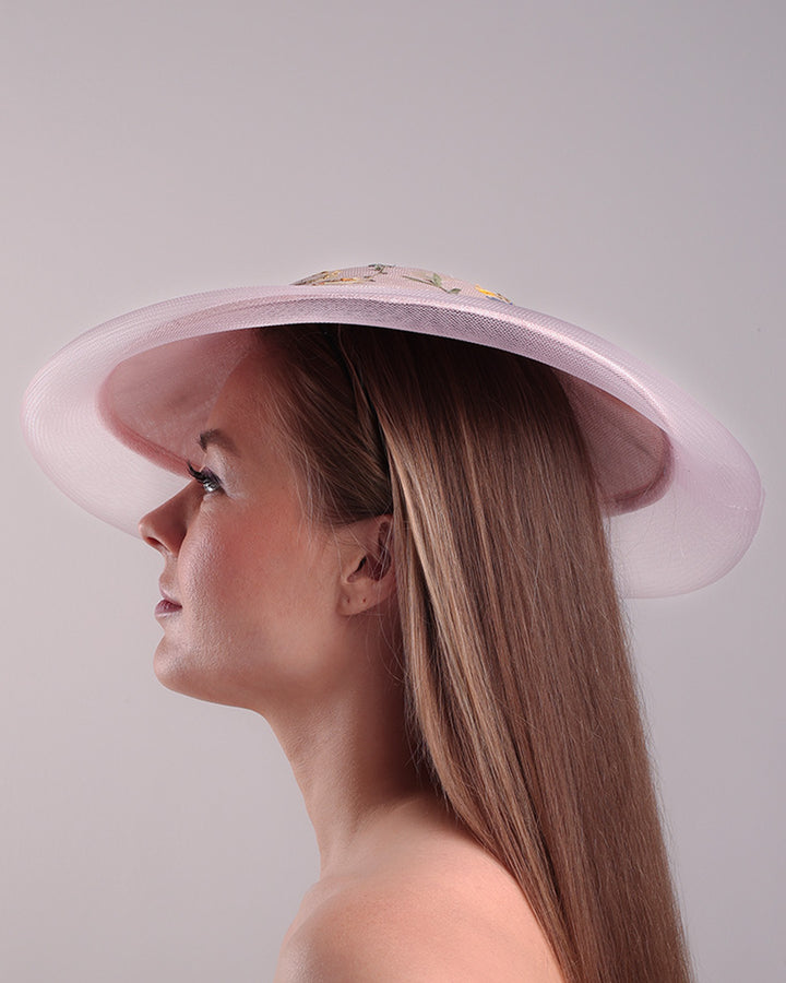 The model is showcasing the left side of the circle shaped pink sinamay saucer hat, worn tilted to the right side. The edges of the circle is embellised with natural pressed dried flowers. The entire cirlce hat extends with pink crinoline at the edge. The saucer hat is fitted with a wire headband and supported with elastic band.
