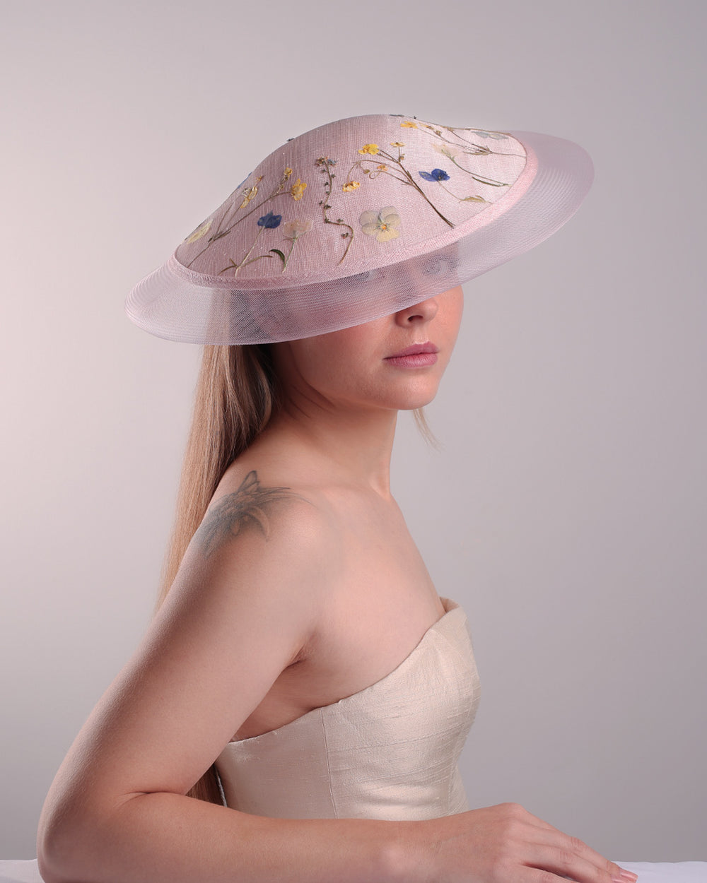 The model is showcasing the front of the circle shaped pink sinamay saucer hat, worn tilted to the right side. The edges of the circle is embellised with natural pressed dried flowers. The entire cirlce hat extends with pink crinoline at the edge. The saucer hat is fitted with a wire headband and supported with elastic band.