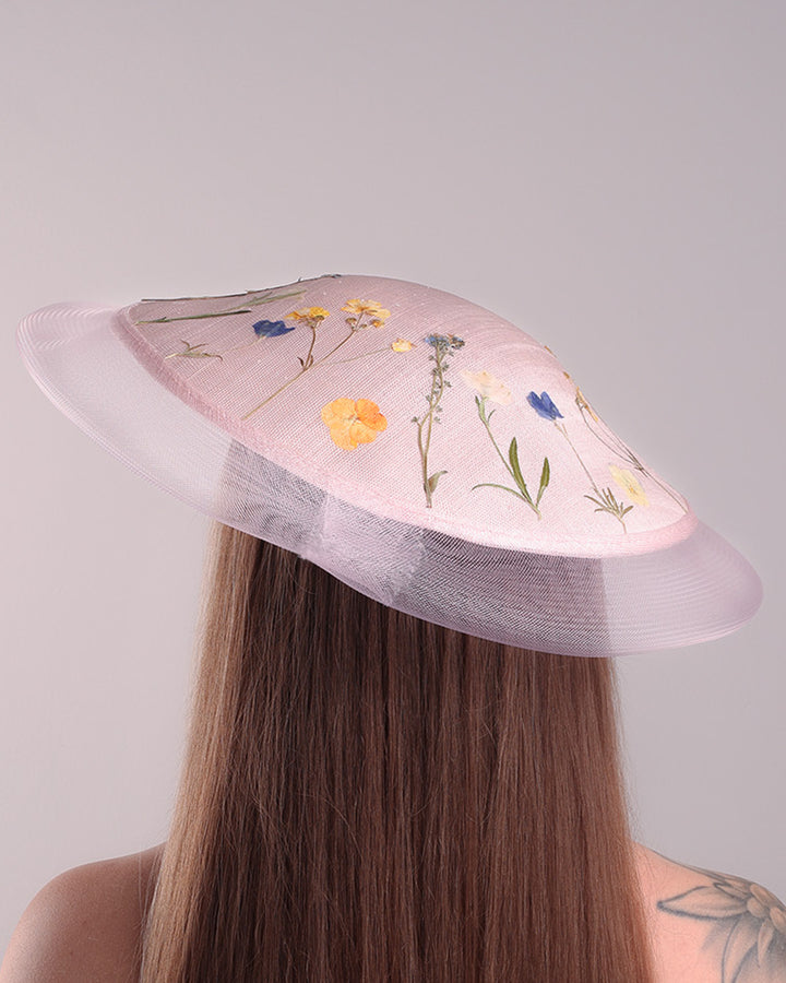 The model is showcasing the back of the circle shaped pink sinamay saucer hat, worn tilted to the right side. The edges of the circle is embellised with natural pressed dried flowers. The entire cirlce hat extends with pink crinoline at the edge. The saucer hat is fitted with a wire headband and supported with elastic band.