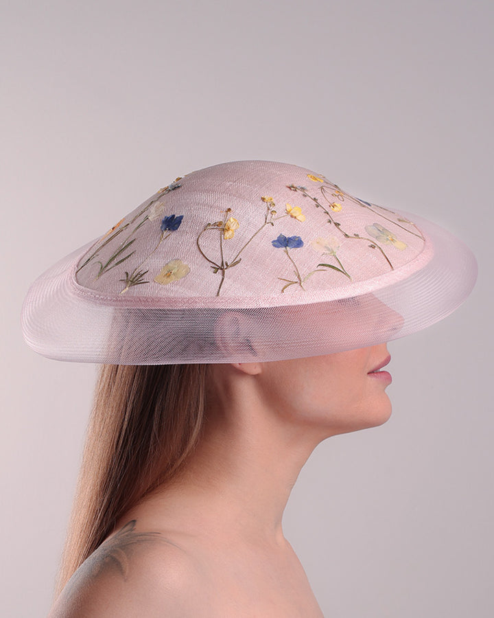 The model is showcasing the right side of the circle shaped pink sinamay saucer hat, worn tilted to the right side. The edges of the circle is embellised with natural pressed dried flowers. The entire cirlce hat extends with pink crinoline at the edge. The saucer hat is fitted with a wire headband and supported with elastic band.