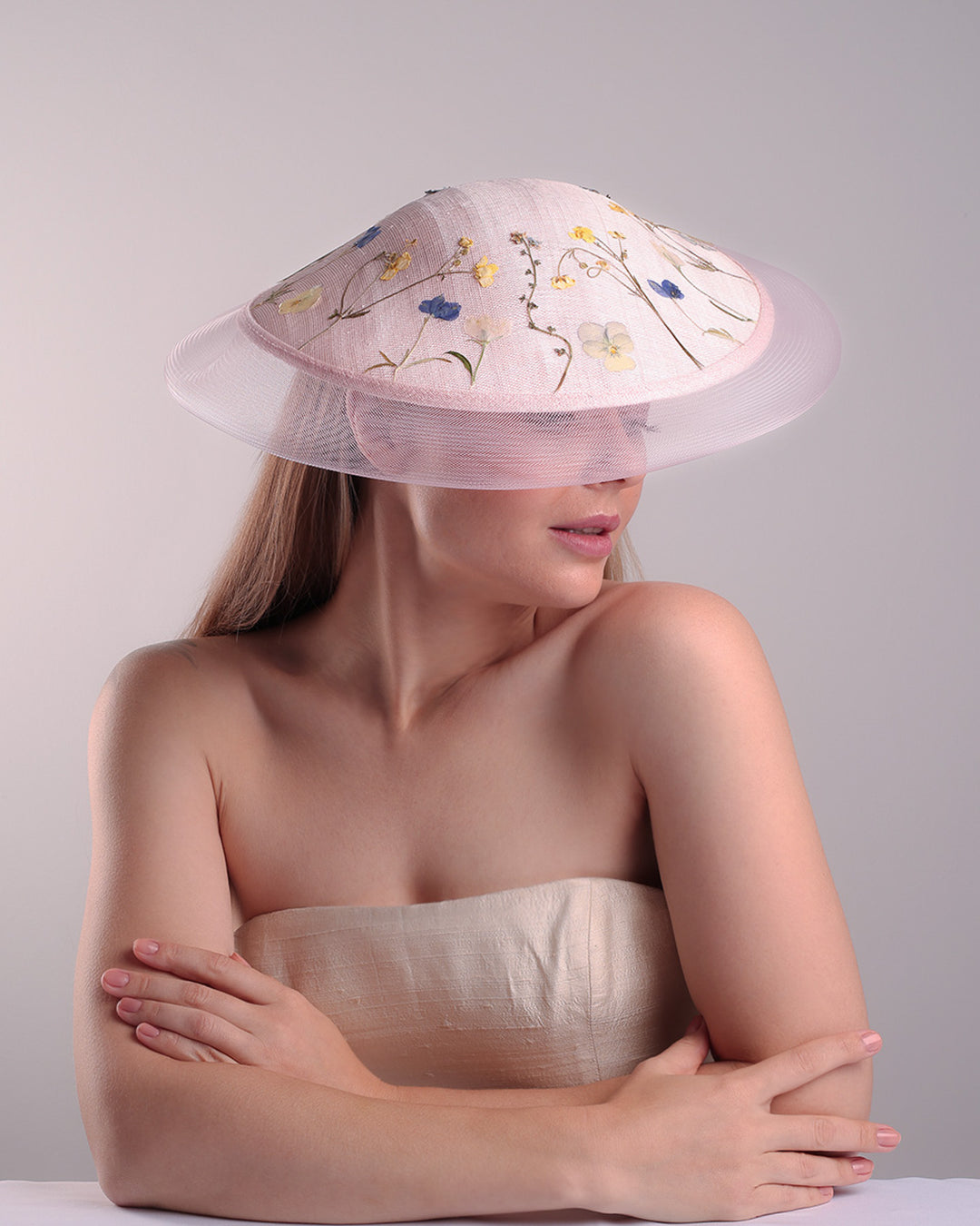 The model is showcasing the front top side of the circle shaped pink sinamay saucer hat, worn tilted to the right side. The edges of the circle is embellised with natural pressed dried flowers. The entire cirlce hat extends with pink crinoline at the edge. The saucer hat is fitted with a wire headband and supported with elastic band.