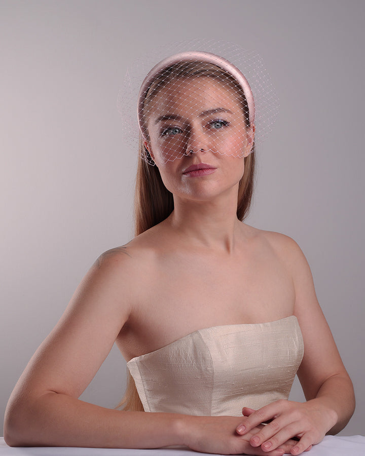 Model is presenting the fornt of a thin slightly high, silk pink headband that is embellished with a pink veil.