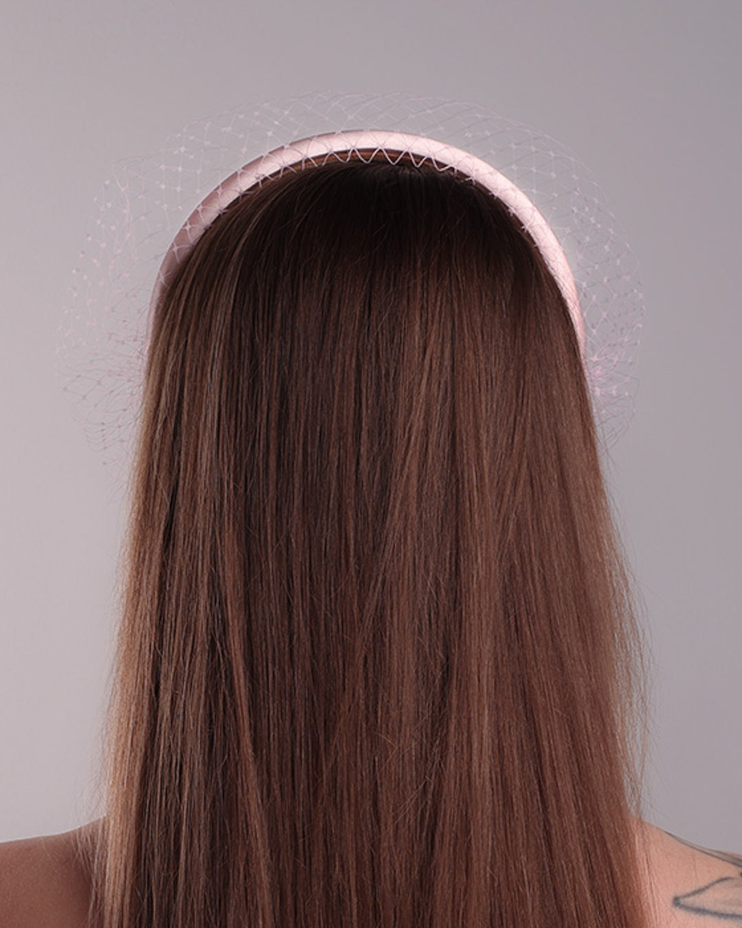 Model is presenting the backside of a thin slightly high, silk pink headband that is embellished with a pink veil.
