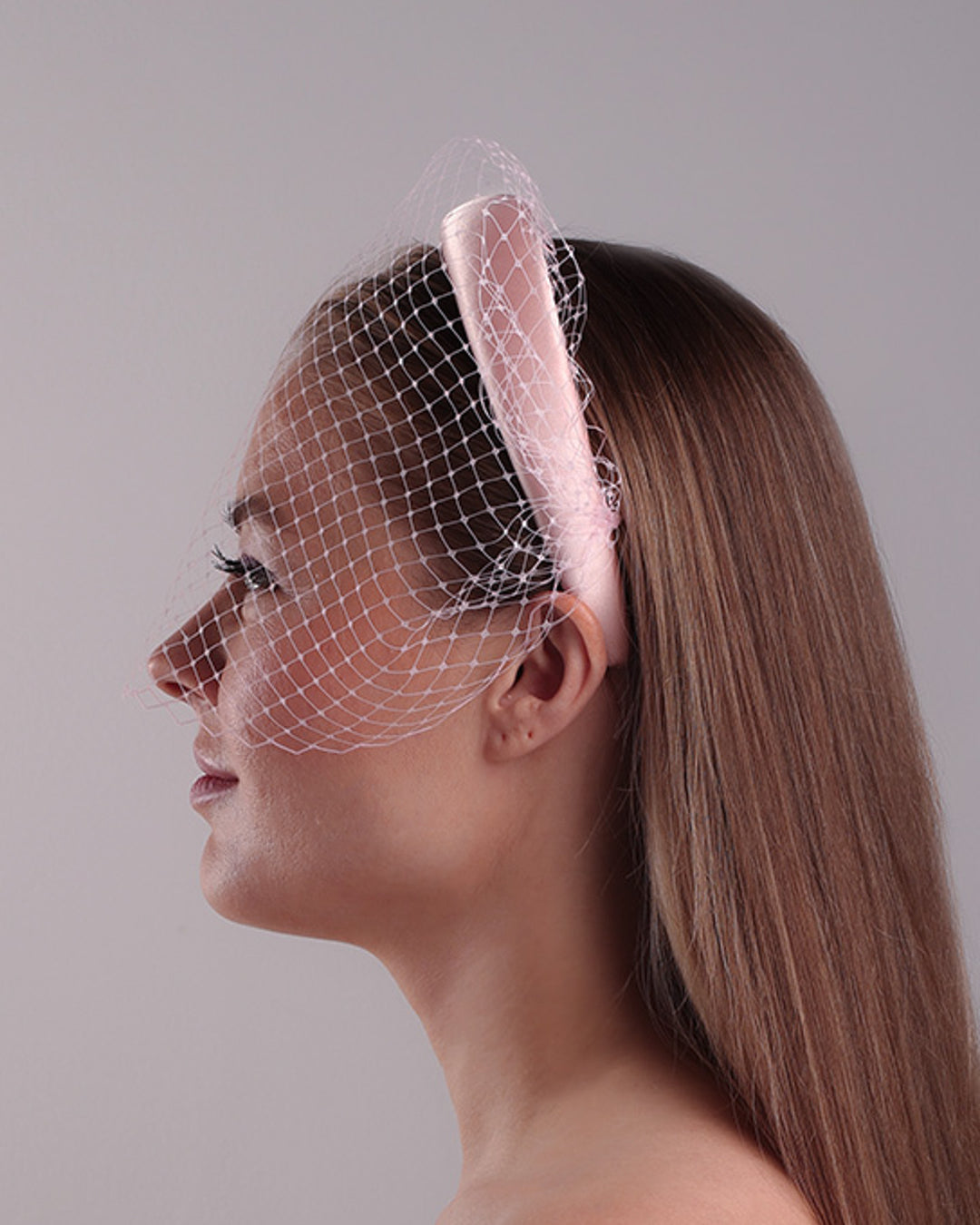 Model is presenting the left side of a thin slightly high, silk pink headband that is embellished with a pink veil.