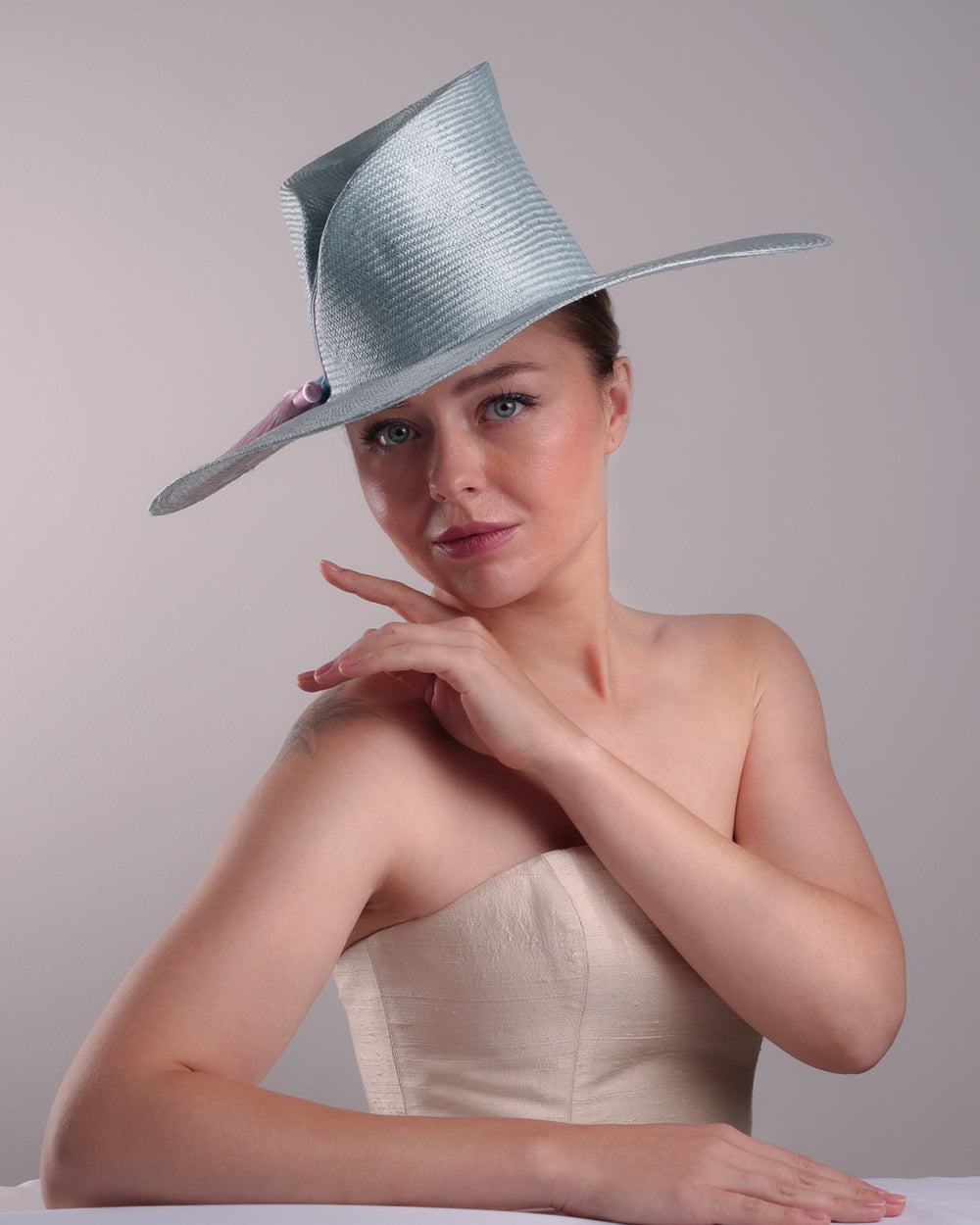 Model is presenting the front of a blue parasisal hat with asymetric crown and wide brim. Decorated with blue and pink tassels.