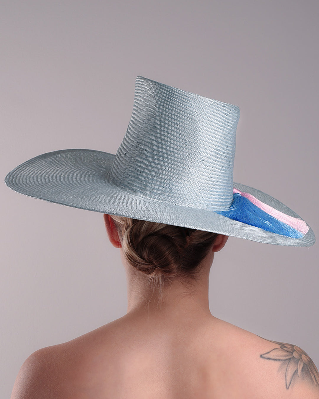 Model is presenting the back of a blue parasisal hat with asymetric crown and wide brim. Decorated with blue and pink tassels.