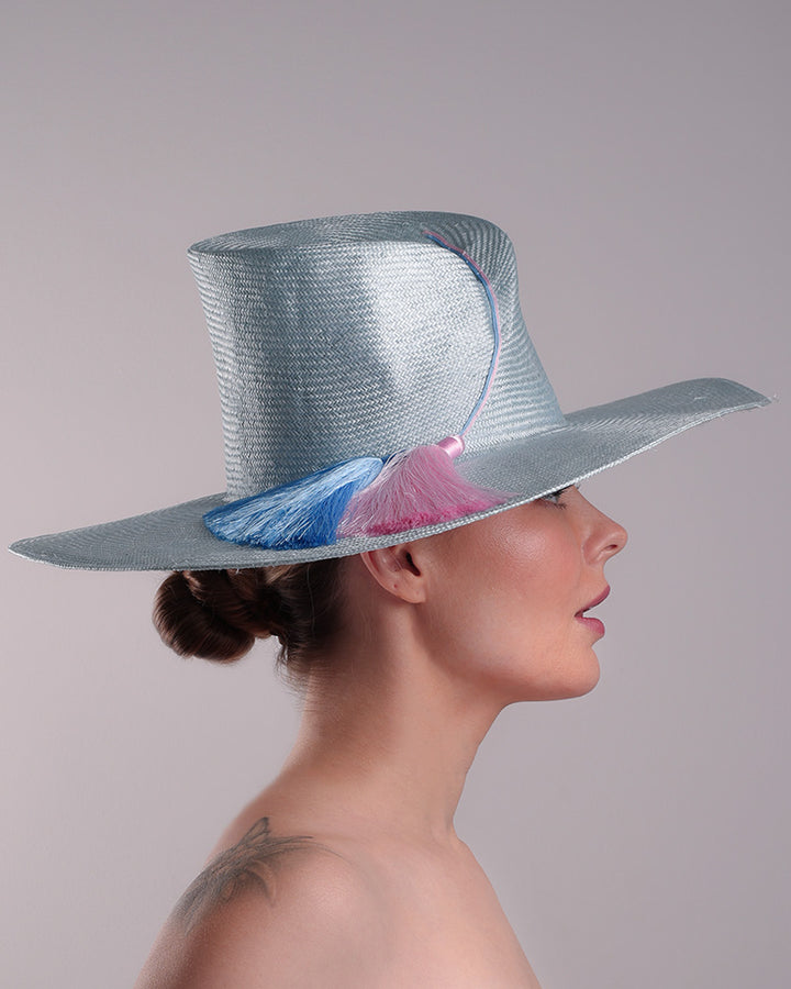 Model is presenting the right side of a blue parasisal hat with asymetric crown and wide brim. Decorated with blue and pink tassels.