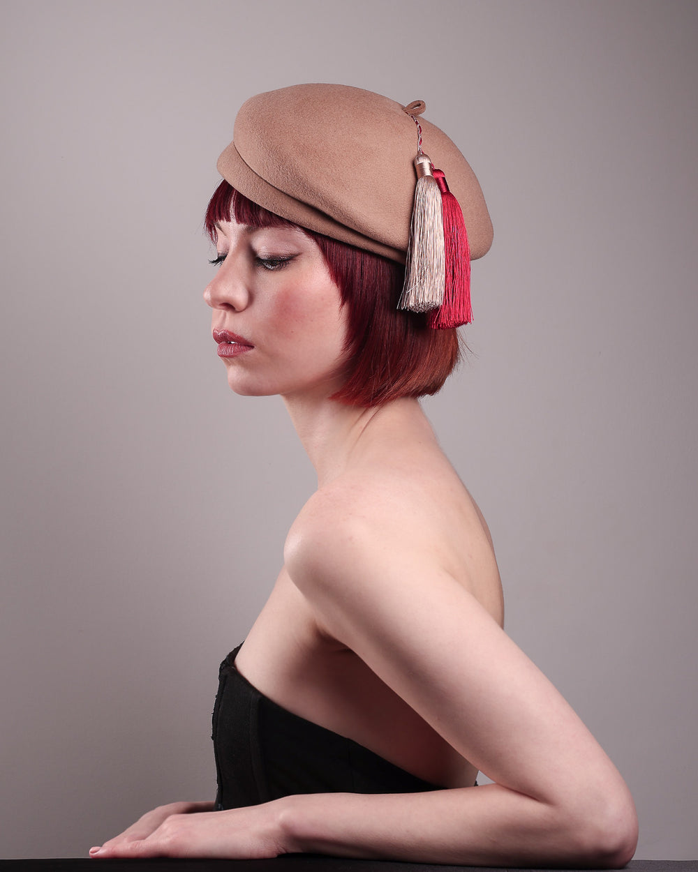The model is showcasing the right angle of the Elera Felt Beret Hat, made of smooth camel wool felt. This structured beret features a dramatic two-tone tassel in crimson and cream, gently draped to one side from a polished metallic accent. Designed by Merve Bayindir. 