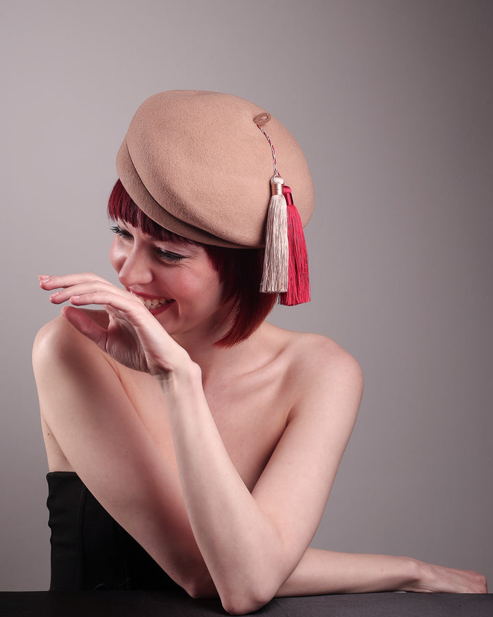 The model is showcasing the right angle top side of the Elera Felt Beret Hat, made of smooth camel wool felt. This structured beret features a dramatic two-tone tassel in crimson and cream, gently draped to one side from a polished metallic accent. Designed by Merve Bayindir. 