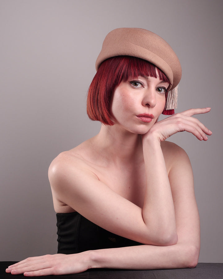 The model is showcasing the left front angle of the Elera Felt Beret Hat, made of smooth camel wool felt. This structured beret features a dramatic two-tone tassel in crimson and cream, gently draped to one side from a polished metallic accent. Designed by Merve Bayindir. 