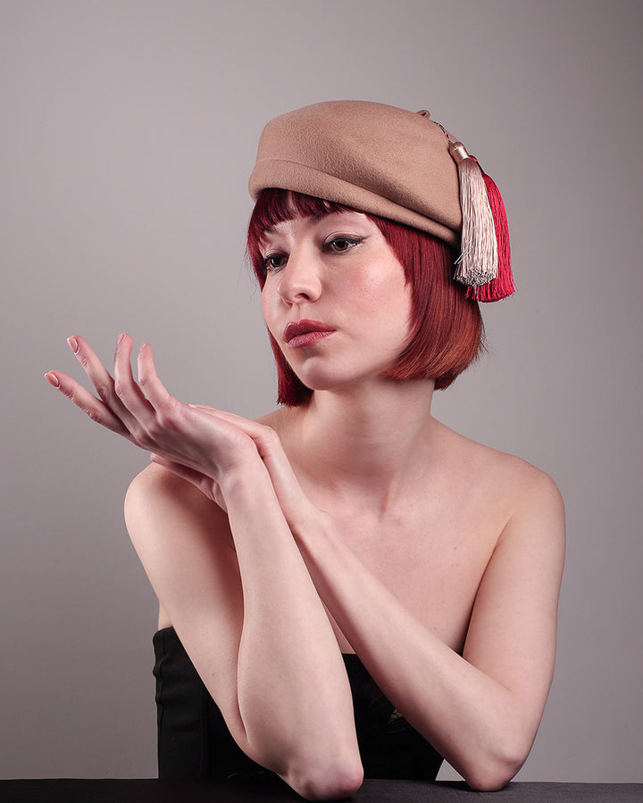 The model is showcasing the right below angle of the Elera Felt Beret Hat, made of smooth camel wool felt. This structured beret features a dramatic two-tone tassel in crimson and cream, gently draped to one side from a polished metallic accent. Designed by Merve Bayindir. 