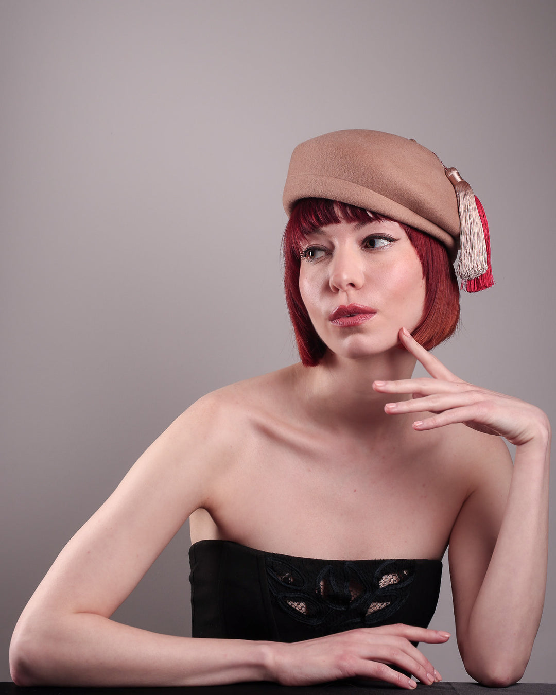 The model is showcasing the rigth front side of the  Elera Felt Beret Hat, made of smooth camel wool felt. This structured beret features a dramatic two-tone tassel in crimson and cream, gently draped to one side from a polished metallic accent. Designed by Merve Bayindir. 